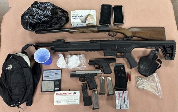 Evidence recovered during a Delaware State Police search warrant, including multiple firearms, handgun magazines, assorted ammunition, suspected narcotics packaged in plastic bags, a digital scale, two cell phones, a backpack, a shoulder holster, a box of sandwich bags, and other drug-related paraphernalia displayed on an evidence table.